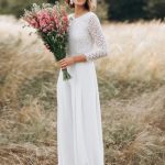 A bride stands in an elegantly lit room, surrounded by soft white curtains and delicate floral arrangements. She is wearing a stunning bridal jumpsuit made from flowing chiffon fabric, which drapes beautifully over her frame. The jumpsuit features a high neckline with subtle lace detailing along the edges, giving it a sophisticated yet modern look. The bodice is form-fitting, while the wide-legged pants create a graceful silhouette that moves fluidly as she stands.